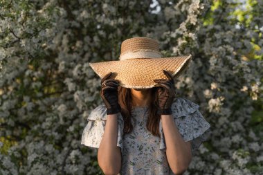 Genç bir kadın yüzünü hasır bir şapkayla örtüyor. Elleri siyah, eski, dantel eldivenli. Bahar çiçeği elma bahçesi. Cottagecore estetiği. Kadınlık pırıl pırıl moda.