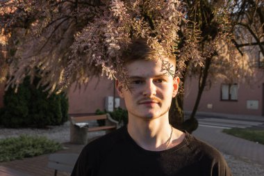 Siyah tişörtlü, yakışıklı, beyaz, sarışın bir adamın portresi. Saç stili, pembe tamariks ağacı dalları. Işık ve gölgeler. Doğayla uyum..