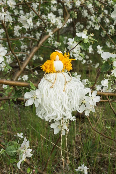 Handmade faceless doll knitted from white and yellow threads in spring blossom apple tree garden. Macrame technique. Eco decoration for the interior in boho style. Ukrainian nation amulet.