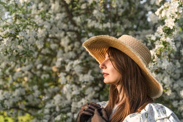 Portrait of attractive caucasian woman in retro straw hat and vintage black lace gloves. Side view. Cottagecore aesthetics. Enjoy spring nature, relax in countryside blossom apple tree garden.