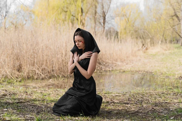 A young widow in a black mourning dress and headscarf kneeling on the ground, praying and crying with bloody tears. Ukrainian woman. Victim of war. Stop war, fighting and genocide in Ukraine.