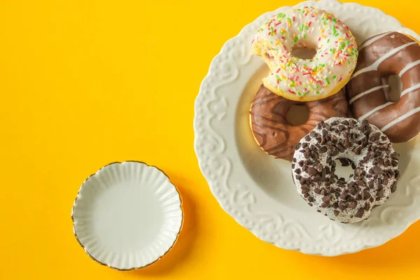 Colorful glazed donuts on a vintage white plate isolated on the yellow background. Pile of assorted donuts with empty round vintage plate near. Top view. Unhealthy eating concept. National Donut Day.