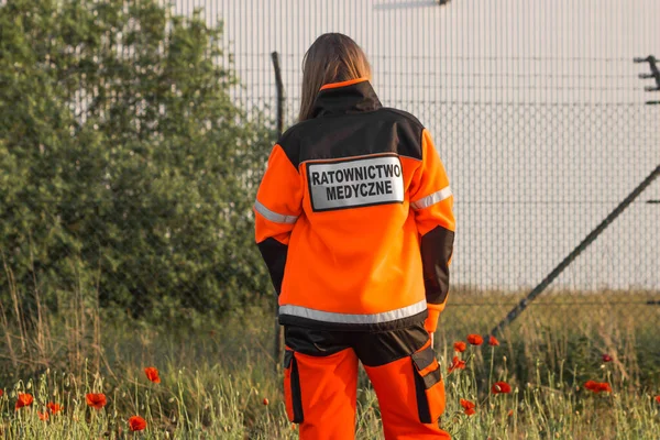 Woman a polish ambulance worker standing back in high visibility medical orange uniform with inscription Ratownictwo Medyczne - Emergency medical Services. Successful technician or doctor in Poland.