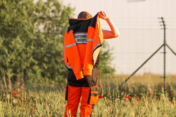 Woman a polish ambulance worker standing back in medical orange uniform with inscription Ratownictwo Medyczne - Emergency medical Services. Puts on a jacket. Successful technician or doctor in Poland.