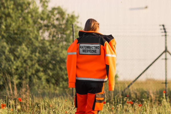 Woman a polish ambulance worker standing back in medical orange uniform with inscription Ratownictwo Medyczne - Emergency medical Services. Puts on a jacket. Successful technician or doctor in Poland.