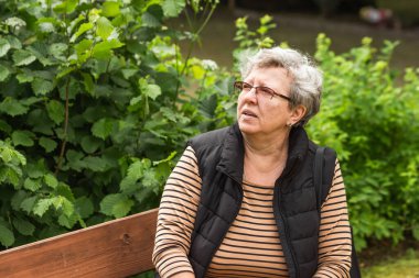 Gözlüklü, parktaki bankta oturup dinlenen düşünceli yaşlı bir kadının portresi. Gri saçlı kadın boş zamanlarını yeşil yaz parkında geçiriyor. Yaşlı yaşam konsepti.