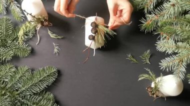 Top view of florist hands making a christmas decor for candles with fir branch, pine cones and thread twine in rustic style. Black background. Eco decor for winter holidays. DIY and handicraft concept