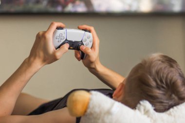 Poznan, Polonya - Ağustos 2022. Evdeki bulanık TV ekranının önünde Play Station 5 Dual Sense kumandasıyla oynayan bir adam. Modern joystick 'i tutan ve video oyunları oynayan insanların elleri. Hafta sonu etkinliği.