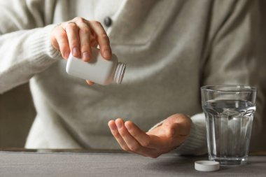 Close-up mans hands holds white plastic bottle with pills and ready to take supplement pill with glass of water. Ill man taking medication. Sick person need medicine against a headache and cold.
