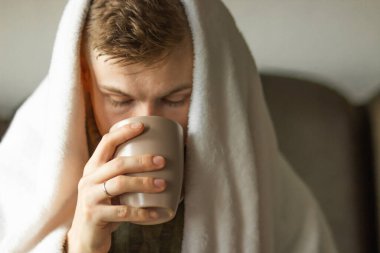 Caucasian young man has fever and common cold sitting at home, wrapped in warm blanket and drinking a cup of antipyretic. Sick day at home concept. Autumn morning scene. Portrait of unhealthy man.