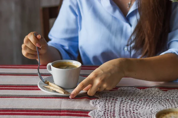 Woman enjoys morning cup of coffee on the table with striped tablecloth in cafe. Womans hand holds a spoon. Cozy autumn morning. Coffee break at the office worker. Caffeine free coffee and wellness.