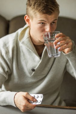 Caucasian young man in white woolen sweater take tablet and drinking clear water from glass on the sofa at home, feels like sick. Ill man taking medication. Prevention of colds. Health care concept. 