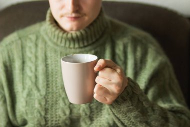 Yeşil örülmüş yün kazak giyen, sıcak kahve ya da çay içen beyaz bir adam. Soğuk sonbaharda ya da kış sabahında elinde kahve fincanı tutan bir adam. Tasarımın için kupa. Isıtma sezonu.