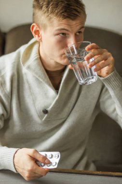 Caucasian young man in white woolen sweater take tablet and drinking clear water from glass on the sofa at home, feels like sick. Ill man taking medication. Prevention of colds. Health care concept. 