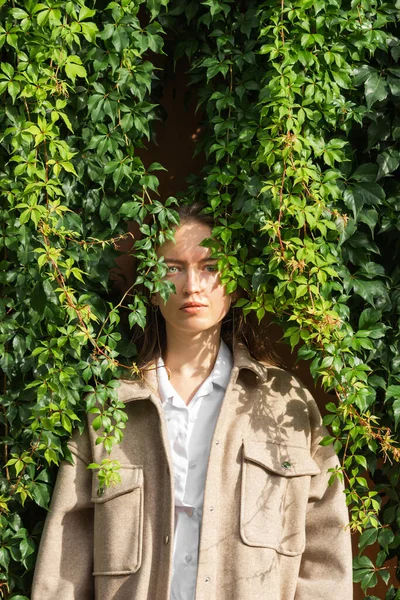 Fashion portrait of caucasian young woman standing next to the eco hedge wall of green wild grape leaves. Female face in shadow patterns of grape leaves. Sunny autumn day. Beauty in nature concept.