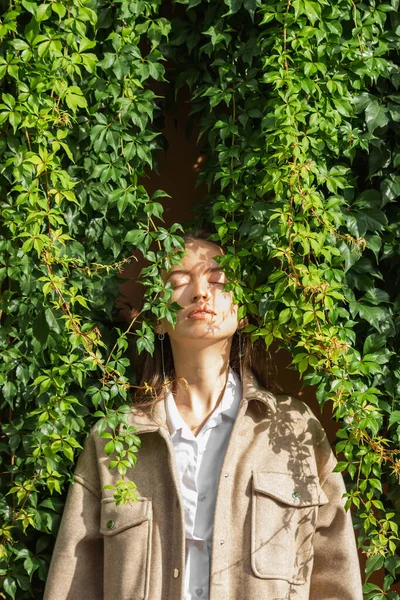 Sensual portrait of caucasian young woman with close eyes standing next to the wall of green wild grape leaves. Female face in shadow patterns of foliage. Sunny autumn day. Beauty in nature concept.