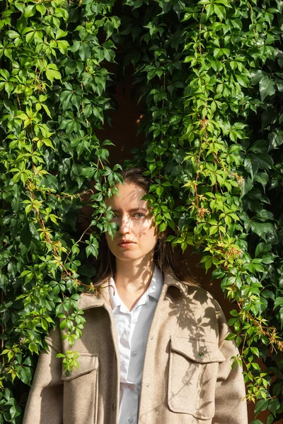 Fashion portrait of caucasian young woman standing next to the eco hedge wall of green wild grape leaves. Female face in shadow patterns of grape leaves. Sunny autumn day. Beauty in nature concept.