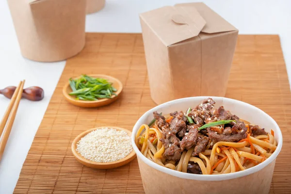 Noodles with meat, peppers, herbs and sesame seeds in a paper disposable plate against the background of a mat with chopsticks and paper boxes for food delivery. Asian street food. Close-up