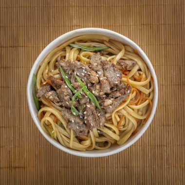 Et, biber, otlar ve susam tohumlu erişteler bir paspasın arka planında tek kullanımlık kağıt bir tabakta. Asya sokak yemeği. Yukarıdan bak. Yakın plan. Kare