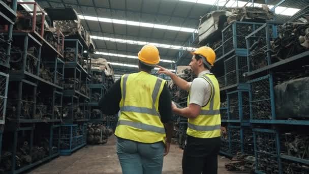 工厂工业中 两名戴安全帽的工人在汽车行业中走来走去 用数字平板检测二手车零部件的库存量 — 图库视频影像