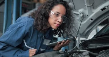 Kadın tamirci, araç motorlarını kontrol eden bir tablet bilgisayar kullanıyor. Uzman, motor bölmesinin içindeki kırık parçaları bulmak için arabayı inceliyor. Araba Servisi.