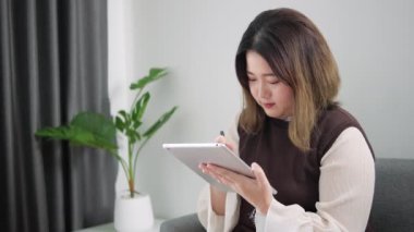 Evdeki oturma odasında dijital tablet kullanan ve stilus kalemle notlar yazan Asyalı kadın, evden çalışıyor..
