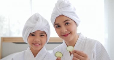 Beyaz bornozlu Asyalı anne ve kız gözlerinde salatalıkla eğleniyorlar. Kızlar için güzellik merkezi prosedürleri. Aile ve Güzellik Konsepti.