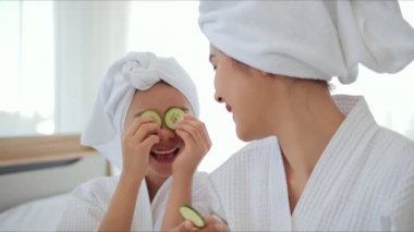 Beyaz bornozlu mutlu Asyalı anne ve kız gözlerine salatalık sürüp eğleniyorlar. Kızlar için güzellik merkezi prosedürleri. Aile ve Güzellik Konsepti.