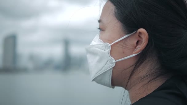 暗い雲が迫り来る都市のスカイラインのビューに対して真剣に表現思考と屋外に立って顔マスクを身に着けている疲れアジアの女性のクローズアップ — ストック動画