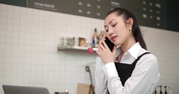 カフェで働くアジア人女性コーヒーショップの従業員バリスタ 笑顔の女性ウエイトレスのレジ係が電話で顧客から注文コーヒーとパン屋を取っています 小規模事業主とパートタイムの仕事のコンセプト — ストック動画
