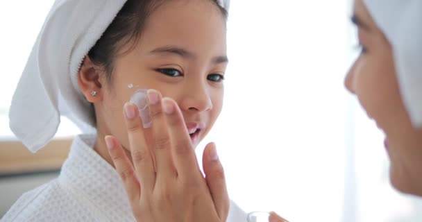 在家里 亚洲妈妈在小女孩的脸颊上加了些治疗霜 她穿着水疗衣 头戴白毛巾躺在床上 — 图库视频影像