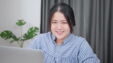 Ev ofisinde çalışan Asyalı kadın bilgisayarı kullanarak kameraya gülümsüyor..