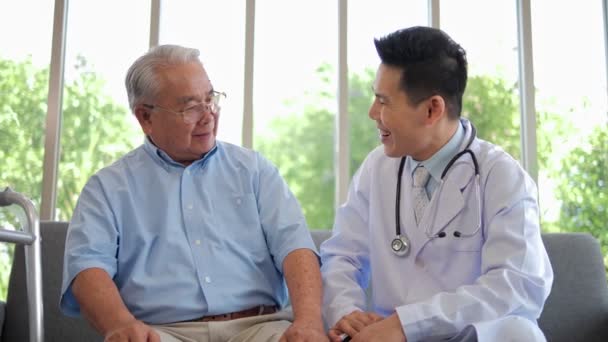 幸せなアジアの男性看護師は 在宅医療訪問で笑顔と笑顔のおじいちゃんをサポートする医療サービスを提供し 医師は共感を与える自宅病院でソファに座って引退した患者を奨励 — ストック動画
