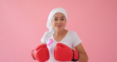 Pembe kurdeleli ve boks eldivenli Asyalı kadın pembe arka plandaki kameraya bakıyor. Göğüs kanseri kavramı.
