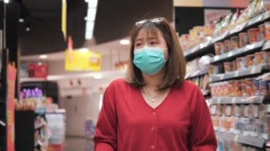 Yüz maskesi takan Asyalı kadın süpermarkette alışveriş arabasını itiyor. Coronavirüs krizi sırasında ya da covid19 salgını sırasında raftan alışveriş malzemesi almak isteyen bir kız..