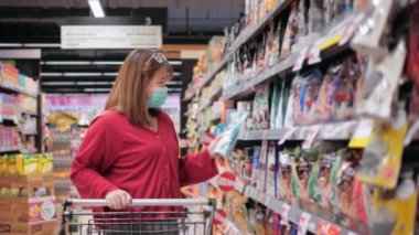 Yüz maskesi takan Asyalı kadın süpermarkette alışveriş arabasını itiyor. Coronavirüs krizi sırasında ya da covid19 salgını sırasında raftan alışveriş malzemesi almak isteyen bir kız..