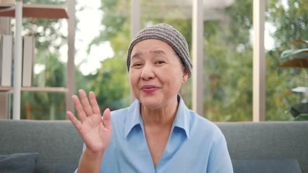 60代のアジア系の高齢者の女性がカメラ会議を自宅で呼び出す話をしている 笑顔シニア成熟した大人の女性祖母ビデオチャット通信オンライン上のソファ ウェブカメラビュー — ストック動画