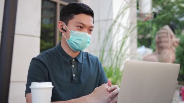在室外露天咖啡馆里 戴着防护口罩的亚洲男人正在用笔记本电脑打电话 在该流行病流行期间在城市开展工作 — 图库视频影像