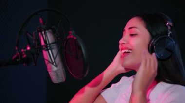 Profesyonel enerjik Asyalı, kulaklık takan kadın şarkıcı, arka planda renkli düşük ışıklarla müzik stüdyosunda kayıt yaparken mikrofonla yeni bir şarkı söylüyor..