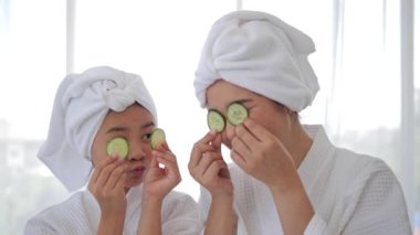 Beyaz bornozlu Asyalı anne ve kız gözlerinde salatalıkla kameraya bakıyorlar. Kızlar için güzellik merkezi prosedürleri. Cilt bakımı, hidratlaştırma ve nemlendirme konsepti.