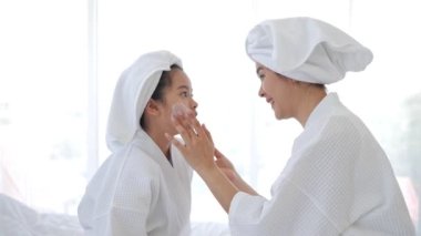 Anne, duştan sonra yatak odasında dinlenirken kızının yüzüne leke kremi sürüyor. Aile güzelliği tedavisi. Gençlik cilt sorunları.