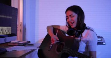 Asyalı bayan müzisyen, evinde neon ışıkla kayıt stüdyosunda oturuyor. Gitar çalıyor ve nota ile şarkı sözü yazıyor.