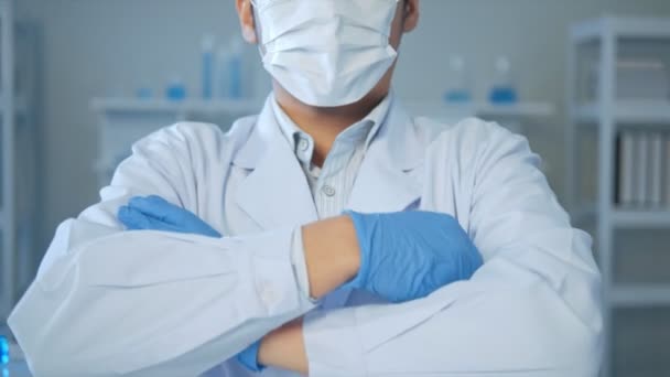 顔のマスクのアジアの男性医師の科学者は 青色の液体をテストする実験室のチューブ内のカメラを見て腕を越えました 医療科学ヘルスケアの概念 — ストック動画