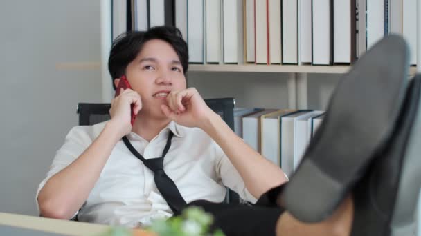 若いアジアのビジネスマンは 仕事中に休憩を取ります 机の上に脚を置け イヤホンをオンにして携帯電話の通話を受信します 話笑顔笑顔 — ストック動画