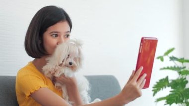 Genç bir kadın tablette video görüşmesi yaparken oturup köpeğini okşuyor. Çekici kız görüntülü konuşma, gülümseme, gülme, evde konuşma..