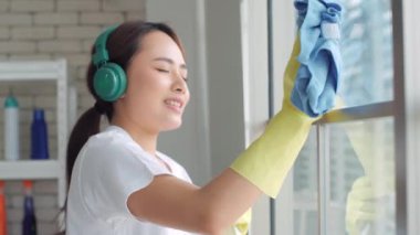 Kulaklıklı genç mutlu Asyalı kadın evde süngerle pencereleri temizlerken müzik dinliyor. Evde temizlik günü.