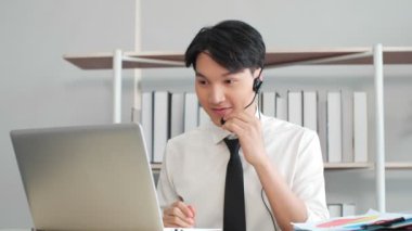 Asian businessman wear wireless headset communicating by conference call speak looking at computer laptop in office, video chat job interview.