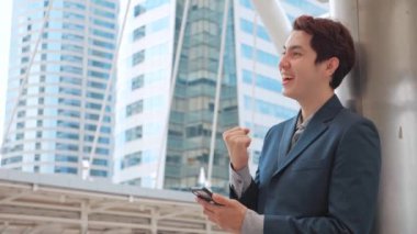 Mutlu Asyalı iş adamı cep telefonuyla iyi haberleri kutluyor modern ofis binasıyla ilgili sürpriz, heyecanlı adam çığlık atıyor evet neşe verici bir başarı cep telefonuna bakıyor.