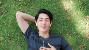 Mutlu genç yakışıklı adam, gözleri kapalı kulaklık takarak rahat yeşil çimenlerde müzik sesi dinliyor. Açılı çekimin üstünde.