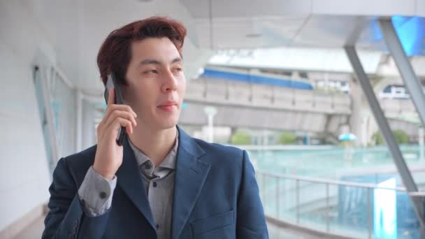 アジアのビジネスマンは 現代的なオフィスビルと携帯電話を話しています 遅い動きの職場で電話を持っている陽気なビジネスマンの閉鎖 幸せなビジネスマンの肖像 屋外電話をかける — ストック動画